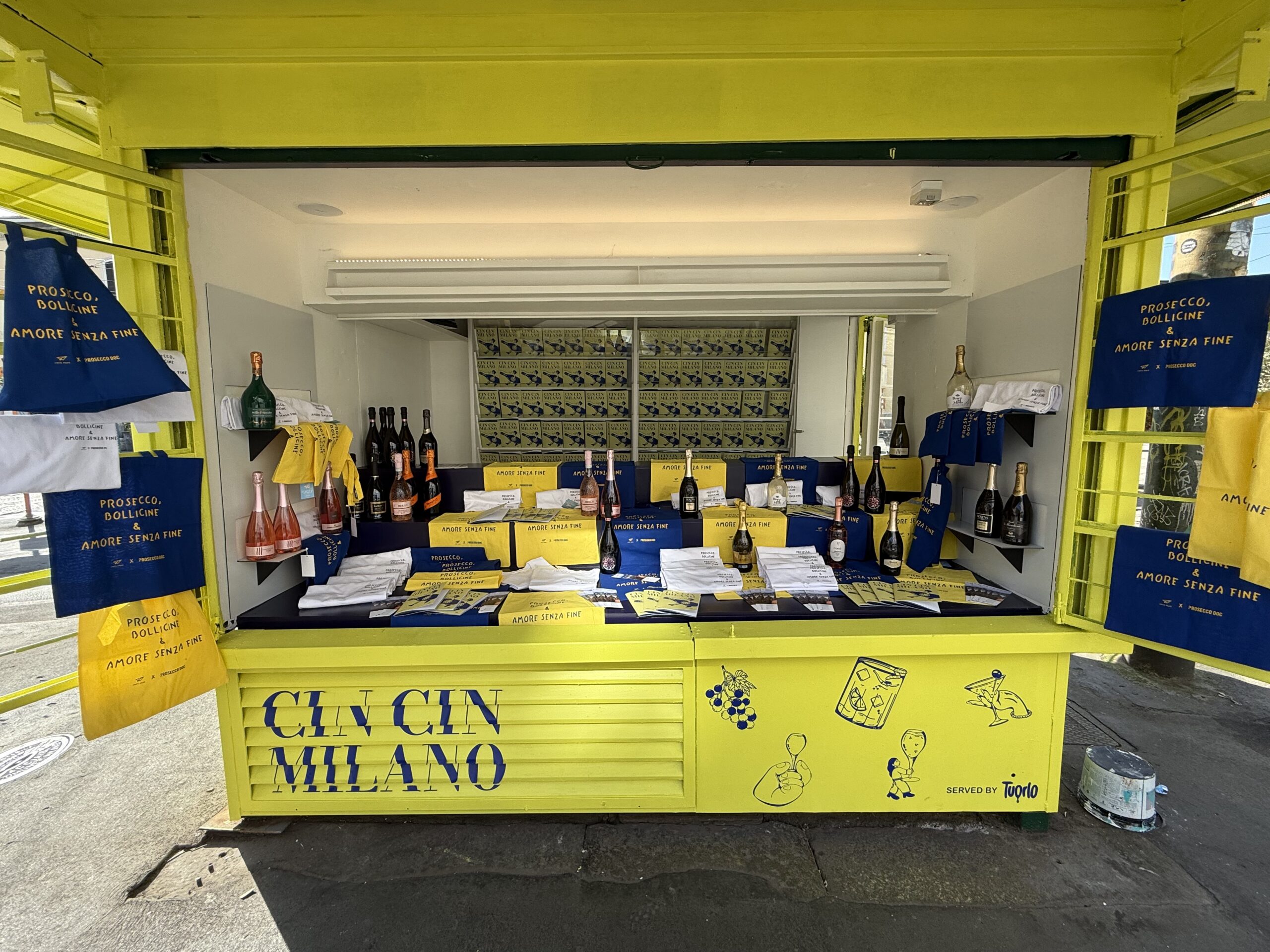 L’installazione 'CIN CIN MILANO' di Prosecco DOC al Fuorisalone 2025: un’edicola pop-up gialla alla fermata metro di Porta Genova, allestita con bottiglie di Prosecco, materiali editoriali, shopper blu e gialle con la scritta 'Prosecco, bollicine & amore senza fine'.