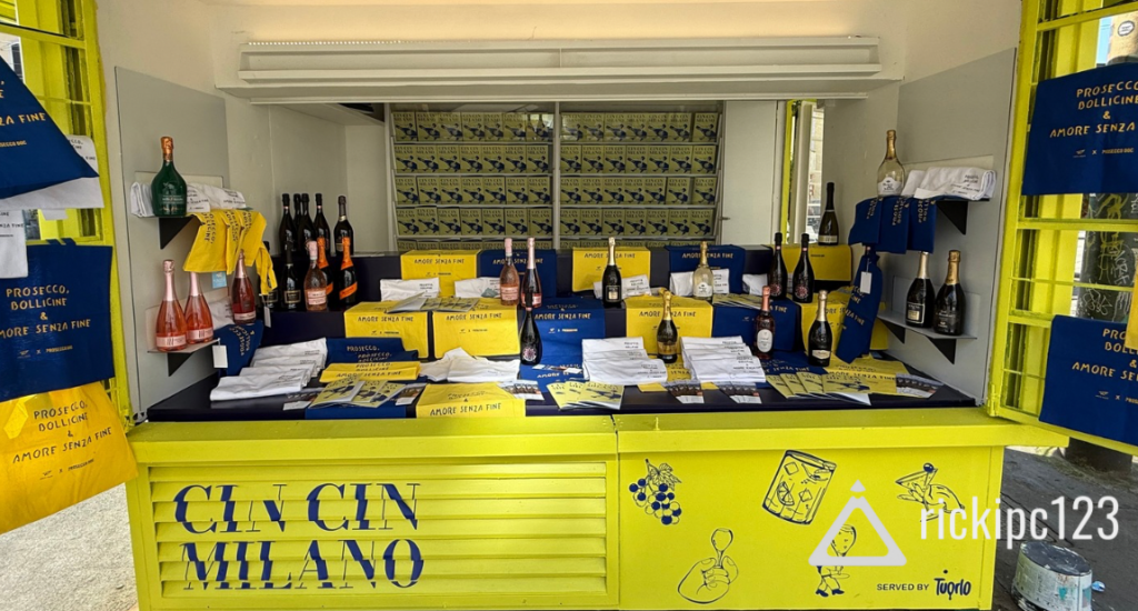 L’installazione “CIN CIN MILANO” di Prosecco DOC al Fuorisalone 2025: edicola pop-up gialla con bottiglie esposte, grafiche blu e gialle, materiali editoriali e shopper con la scritta “Prosecco, bollicine & amore senza fine”.